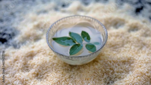 Oryza sativa and Oryza glaberrima or raw lachkari kolam rice grains on a black surface along with a glass bowl containing rice water in it with some rose leaves on top, used in beauty products and fac photo