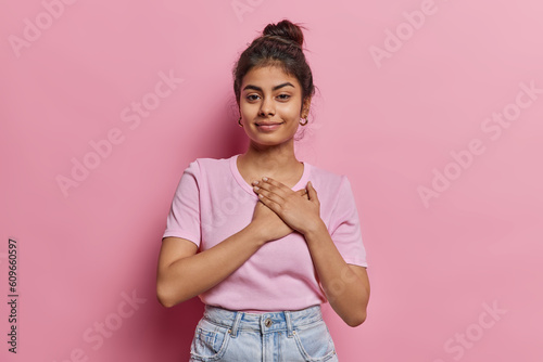 Lovely Indian woman with dark hair gathered in bun gently touches chest her warm expression radiates kindness and affection has eyes filled with compassion and warmth dressed casually stands indoor