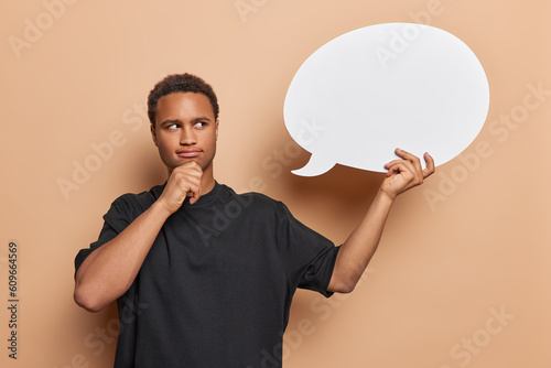 Thoughtful young man holds chin and looks pensively at blank speech bubble considers something what idea to write dressed in casual black t shirt isolated over brown background. Hmm let me think