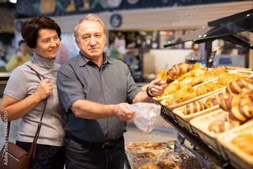 Elderly married couple chooses hot fresh buns and bread at supermarket showcase