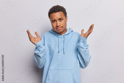 Clueless questioned man spreads palms has unaware expression doesnt know what to do dressed in casual blue hoodie isolated over white background. Hesitant teenager faces difficult choice in life