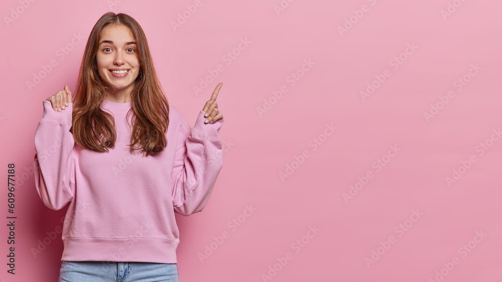 Studio shot of cheerful young European woman cheers over something points index finger above on copy space for your advertising content wears pullover and jeans isolated over pink background Studio shot of cheerful young European woman cheers over something points index finger above on copy space for your advertising content wears pullover and jeans isolated over pink background