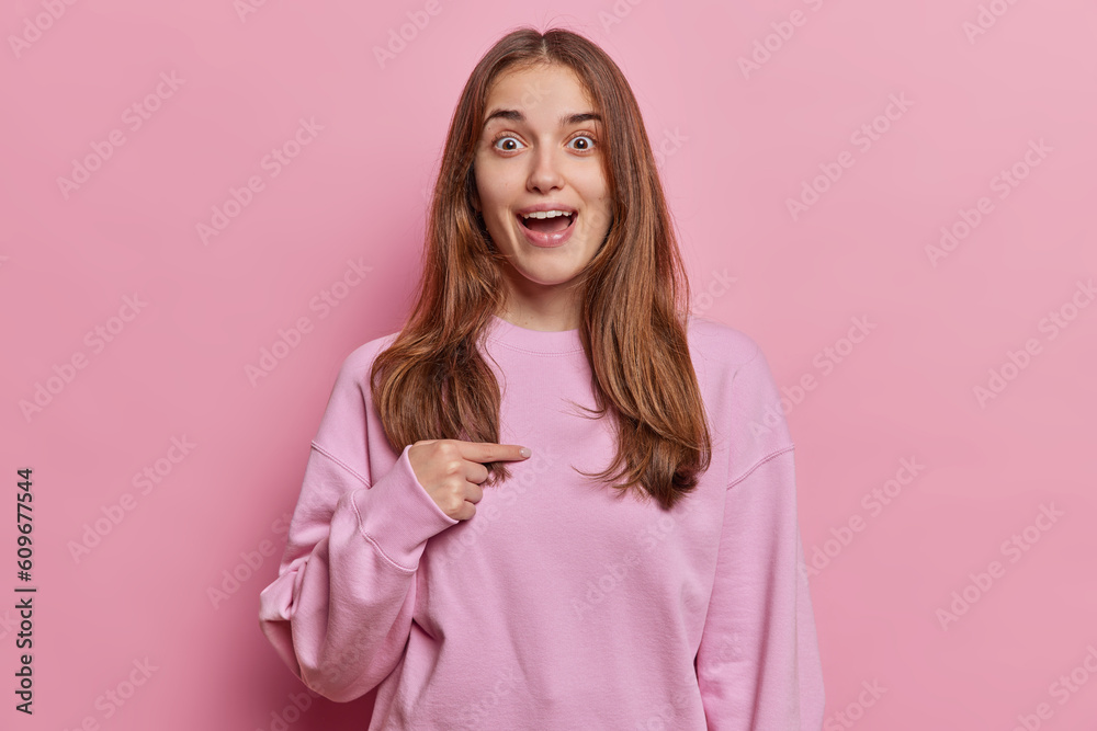 Who me. Surprised young pretty woman points to herself impressed being chosen as appropriate candidate dressed in casual pullover isolated over pink background has eyes wide open from amazement