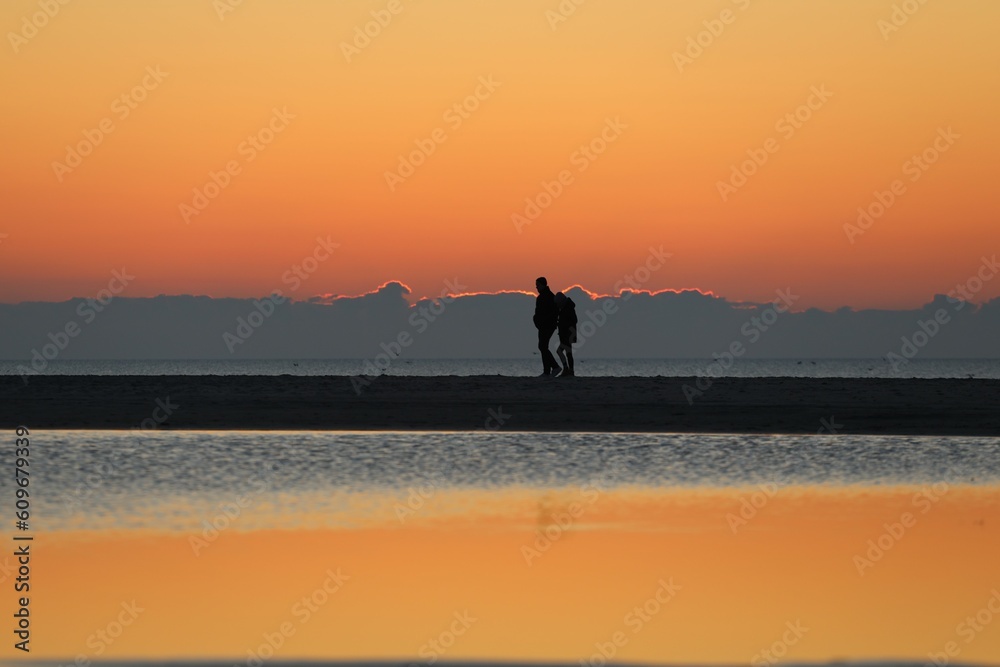 Fototapeta premium Liebespaar geht am Strand entlang im Abendrot hinter Spiegelungen im Wasser