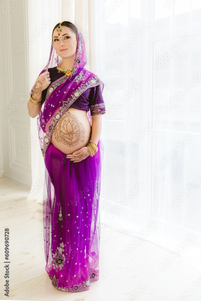 Pregnant woman in purple sari. mehendi. Indian painting on woman's hands and pregnant belly with henna tattoo on white room background Pregnant woman in purple sari. mehendi. Indian painting on woman's hands and pregnant belly with henna tattoo on white room background