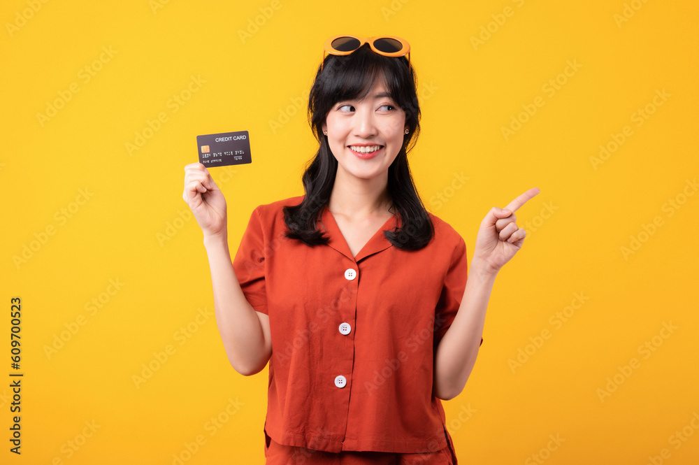 Portrait young beautiful asian woman happy smile dressed in orange clothes showing credit card and point finger to free space isolated on yellow background. Pay and purchase shopping payment concept. Portrait young beautiful asian woman happy smile dressed in orange clothes showing credit card and point finger to free space isolated on yellow background. Pay and purchase shopping payment concept.
