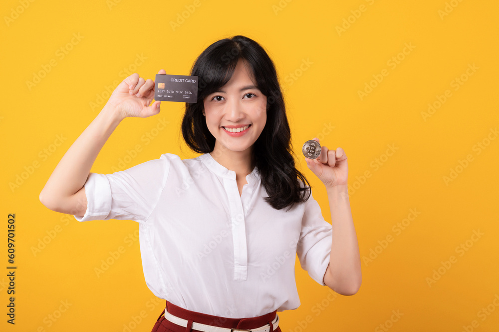 Portrait beautiful young asian woman enterpriser happy smile wearing white shirt and red plants holding credit card and crypto digital currency isolation on yellow background. Wealth concept. Portrait beautiful young asian woman enterpriser happy smile wearing white shirt and red plants holding credit card and crypto digital currency isolation on yellow background. Wealth concept.