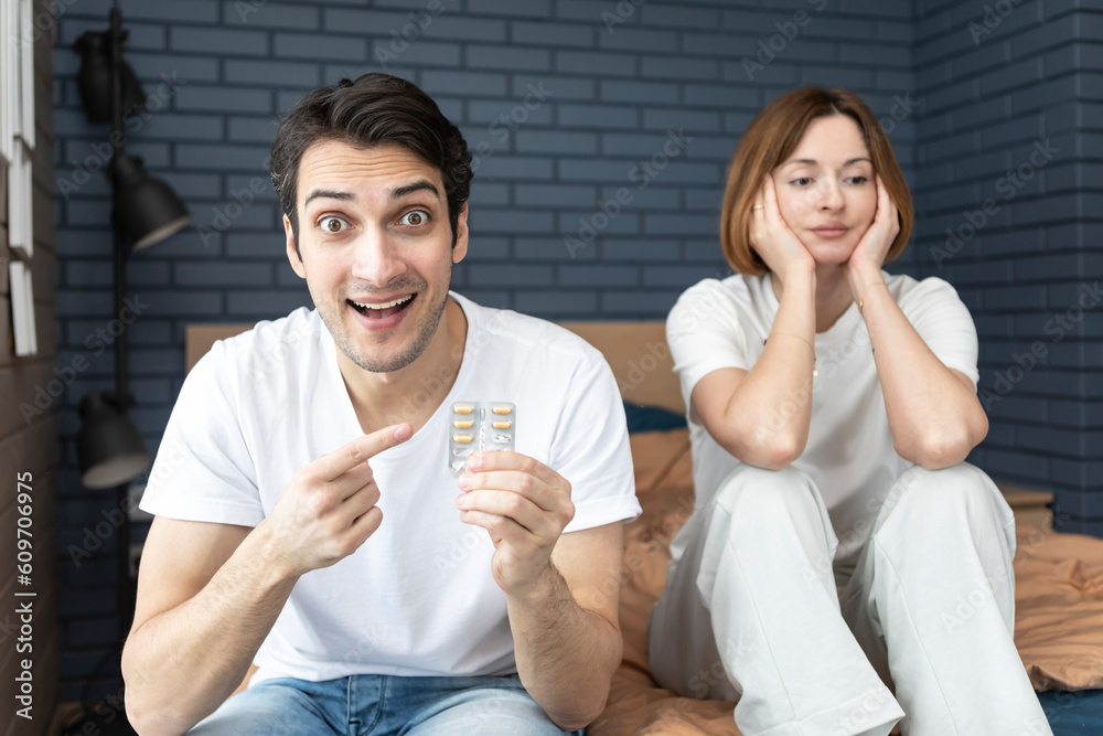 Happy man sitting on the bed, holding tablets and demonstrating his happiness after solving his erectile dysfunction problem Happy man sitting on the bed, holding tablets and demonstrating his happiness after solving his erectile dysfunction problem