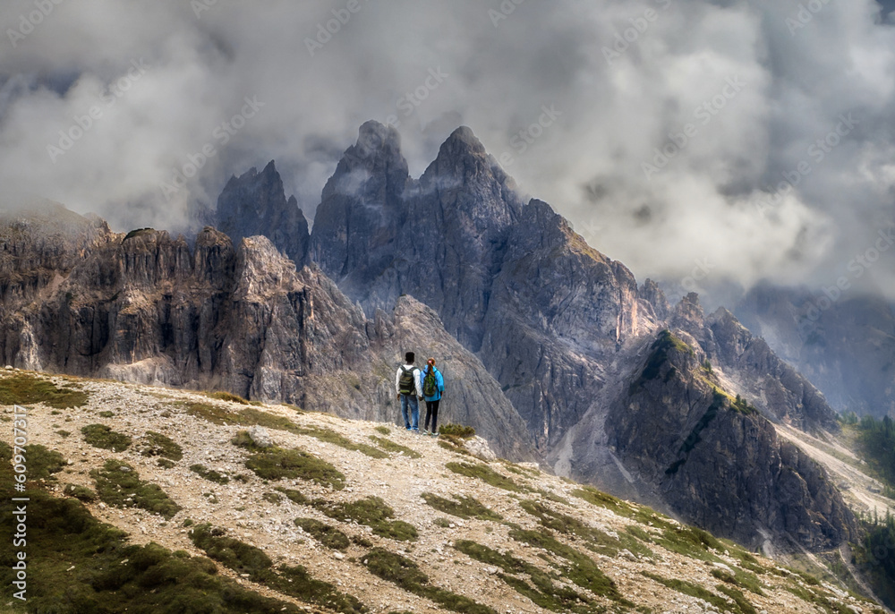 Couple of travelers with backpacks hold hands standing over a cliff and looking at the beautiful mountains landscape. Travel and tourism concept
