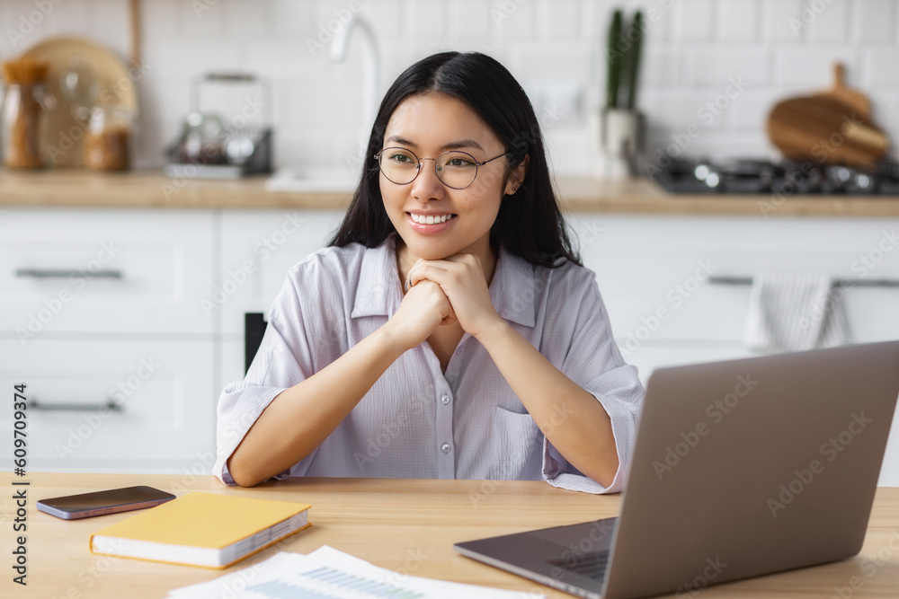 Happy dreamy Asian woman sitting at table with laptop, studying, distance learning or online education. Asian woman freelancer working from home, looking away and smiles friendly Happy dreamy Asian woman sitting at table with laptop, studying, distance learning or online education. Asian woman freelancer working from home, looking away and smiles friendly