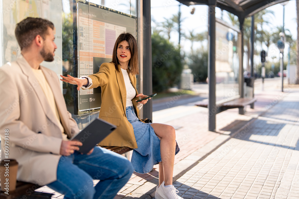 Two business colleagues, beautiful young woman and handsome young man in smart casual clothes sitting together at bus station, communicating, waiting for bus in the city on a sunny day.