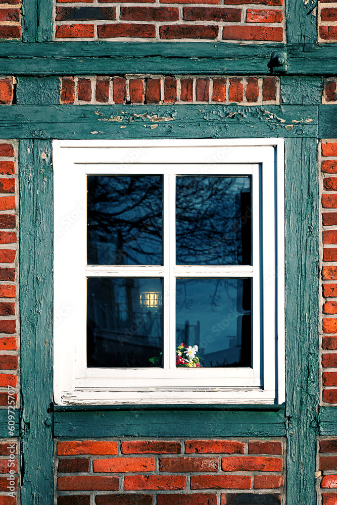 Fototapeta premium Window with a flower pot on the wall of an old half-timbered house in Germany. The branches of a tree, a fence and the light of a lantern are darkly reflected in the window glass.