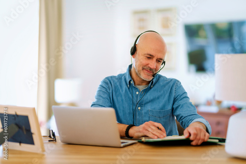 Mid aged man with headset sitting at home and using laptop