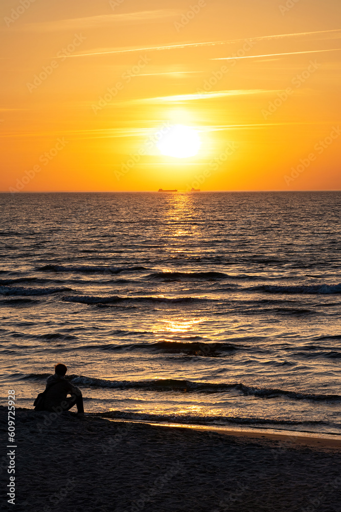 Intense sunset on the sandy beach of the Baltic Sea in Warnemünde in Germany. A person sits on the sand and looks longingly at two distant ships on the horizon.