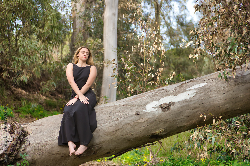 Young woman, blonde and beautiful, with a black dress, sitting on the trunk of a large tree, in the middle of nature, pure and virginal. Concept nature, peace, purity, virginity, trees. Young woman, blonde and beautiful, with a black dress, sitting on the trunk of a large tree, in the middle of nature, pure and virginal. Concept nature, peace, purity, virginity, trees.