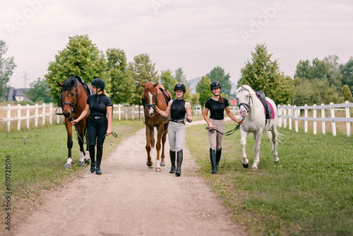 Three beautiful horses with female jockeys, walking side by side along a path near the equestrian farm fields