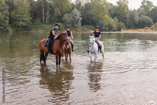Three young women enjoying equestrian leisure, nature, and river, smiling while sitting on horses