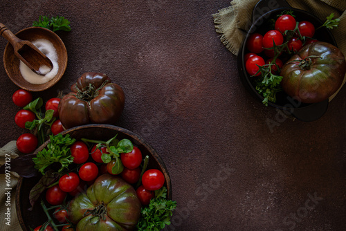 Fresh big tomatoes in dark bowl with cherry tomatoes and green herbs on dark brown background
