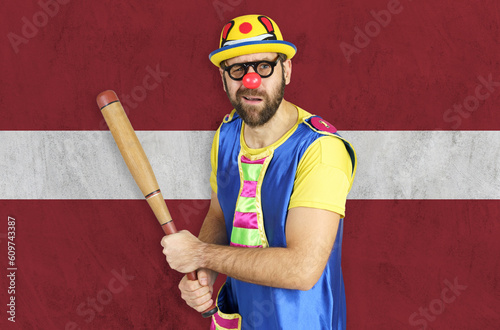 An insecure clown holds a bat in his hands against the background of the flag of Latvia