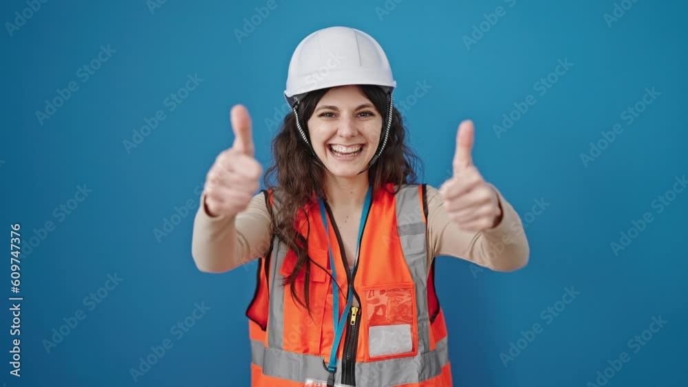 Young beautiful hispanic woman builder smiling confident doing thumbs up gesture over isolated blue background Young beautiful hispanic woman builder smiling confident doing thumbs up gesture over isolated blue background