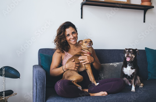 Portrait of cheerful female enjoying leisure time with lovely purebred dogs having fun at comfortable home sofa, Caucasian woman laughing at camera during weekend time with cute mongler doggie