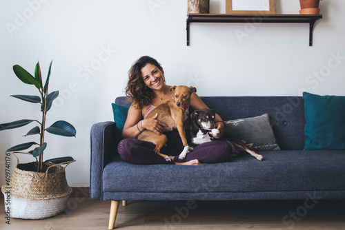 Portrait of happy Caucasian woman in casual clothing hug favourite dogs feeling love emotions during daily routine in home living room, cheerful hipster girl 20s take care of cute pets in collar
