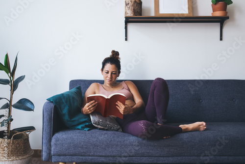 Interested young woman holding literature book and relaxing at home, Caucasian hipster girl reading bestseller with thriller plot resting on comfortable sofa in apartment with stylish flat interior