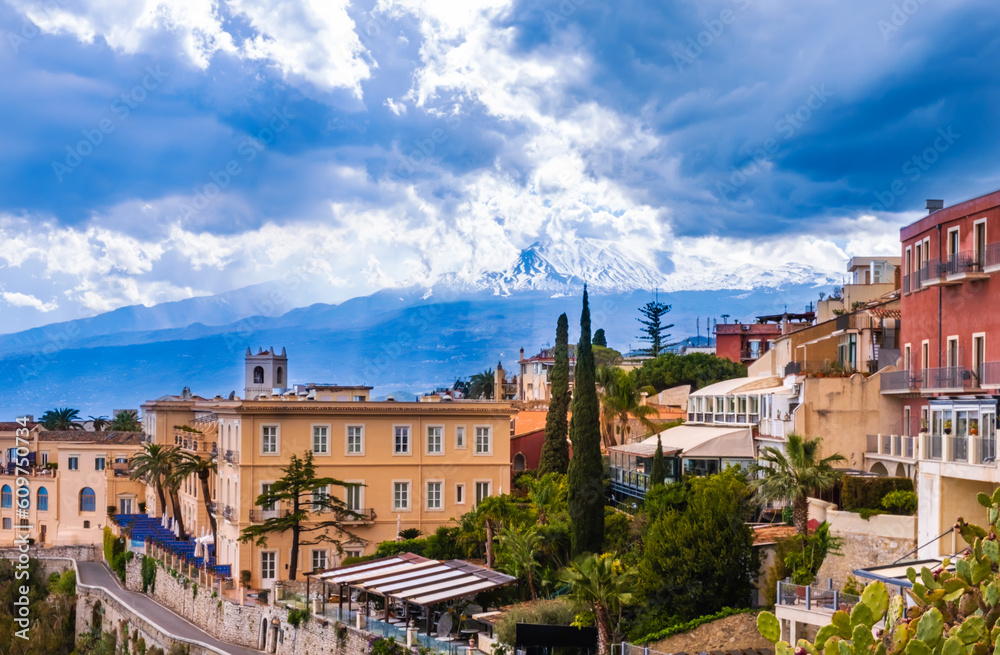 Taormina, Sicily, Italy. Panoramic view over Taormina town on hilltop and Etna mount volcano among clouds in blue sky. Popular tourist destination