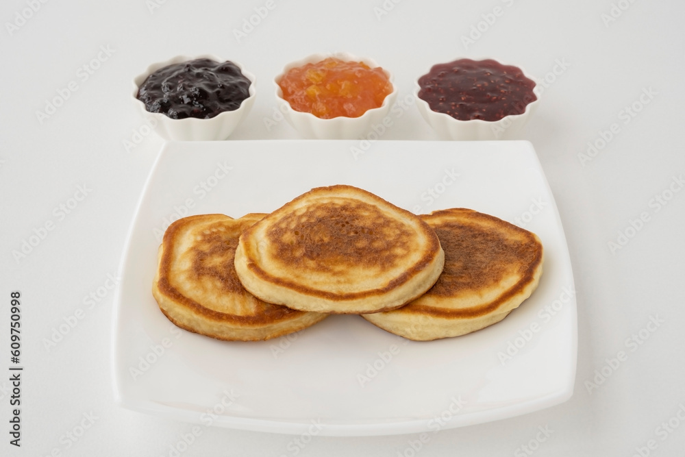 Three pancakes on a white plate and three types of jam in white vases on a white background. Three pancakes on a white plate and three types of jam in white vases on a white background.