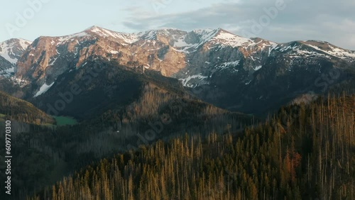 Mountains aerial drone shot snowy mountain peaks green hills covered with pine trees lit with warm sunrise light, epic mountain range view from above, spring season early morning landscape golden hour