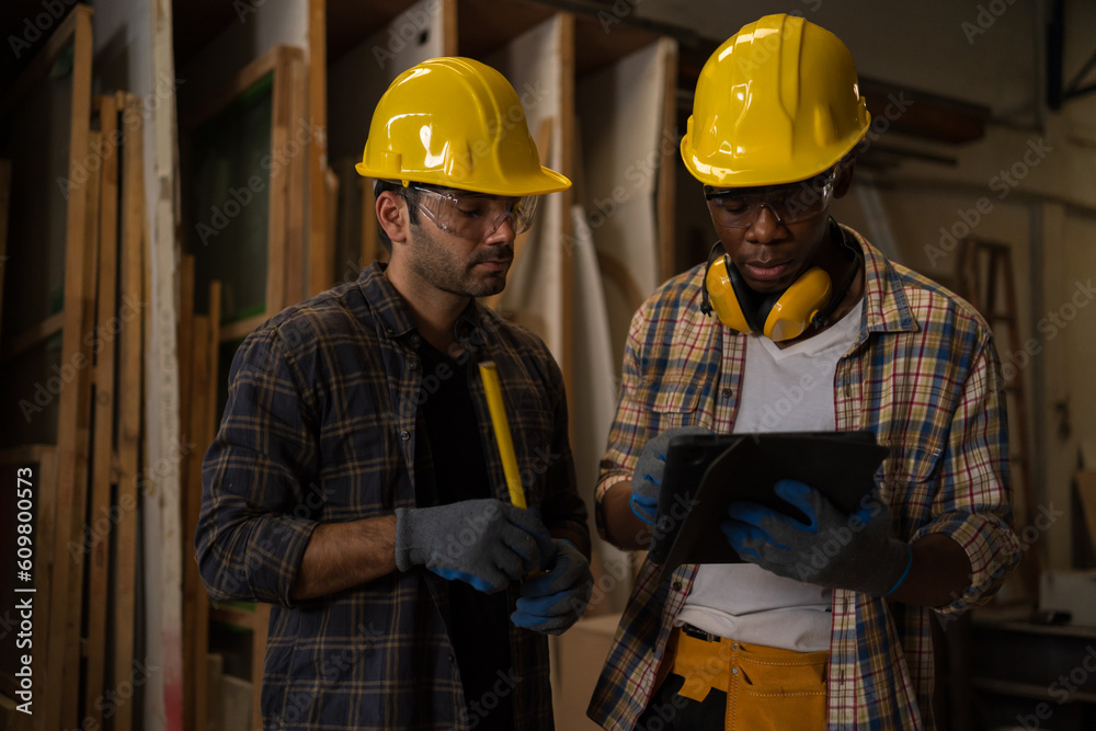 Two diversity professional craftsman or carpenters wear safety equipment. They're talking about woodworking and looking at models from tablets or laptops in Carpenter and wood craftsmanship concept.