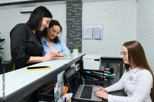 Female doctor and patient discussing medical treatment needs to perform, filling out and signing forms, standing at the reception desk, while the manager receptionist works on laptop in medical clinic