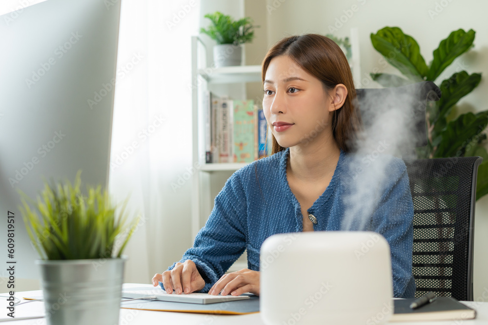 Modern air humidifier device during sitting workplace, happy asian young woman using computer work on internet, enjoying aromatherapy steam scent from essential oil diffuser in room at home office. Modern air humidifier device during sitting workplace, happy asian young woman using computer work on internet, enjoying aromatherapy steam scent from essential oil diffuser in room at home office.