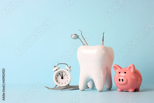 Tooth model with medical instruments, piggy bank and alarm clock on blue background. Investing in dental health care. © WindyNight