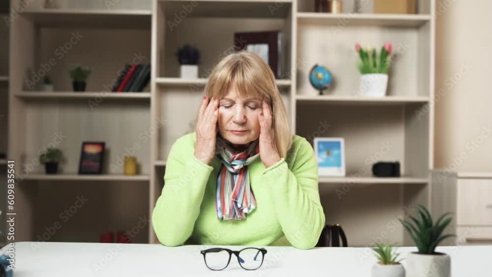 The woman is sitting at the table, she is elderly. She takes off her glasses and places them on the table. The woman also touches her head, indicating that she has a headache. She appears tired. The woman is sitting at the table, she is elderly. She takes off her glasses and places them on the table. The woman also touches her head, indicating that she has a headache. She appears tired.