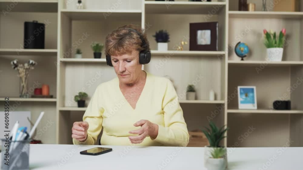 Seated at the table, the elderly woman wears headphones, engrossed in the music playing. She hums along displaying her cheerful disposition. In the backdrop her shelves are adorned with a decorations. Seated at the table, the elderly woman wears headphones, engrossed in the music playing. She hums along displaying her cheerful disposition. In the backdrop her shelves are adorned with a decorations.