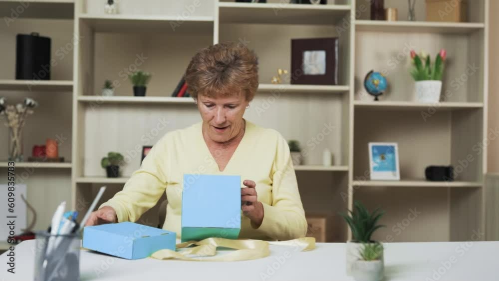 Seated at the table, an elderly woman receives a gift presented to her in a blue box adorned with a golden ribbon. With a joyful expression and a beaming smile, she eagerly unwraps the present Seated at the table, an elderly woman receives a gift presented to her in a blue box adorned with a golden ribbon. With a joyful expression and a beaming smile, she eagerly unwraps the present