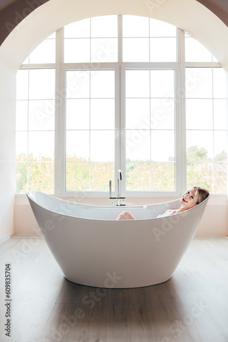 Young beautiful sexy woman having fun while lying in bathtub full of foam at home. Smiling model relaxing in luxury bath interior in front of panorama window. Female enjoying beauty and skincare day © halayalex