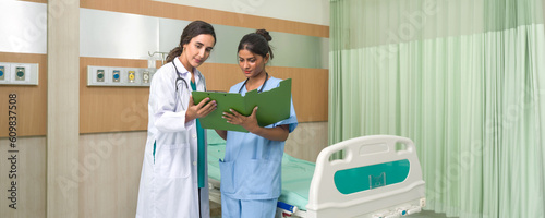 Female doctor in white gown and stethoscope looking at patient data in document folder. Young nurse in blue uniform informed the doctor of the patient examination schedule.