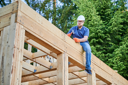 Carpenter constructing double-story wooden-framed house beside the woodland. Bearded man with glasses using hammer to drive nail while wearing safety helmet. Idea of contemporary ecological building.