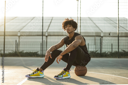 Confident sportsman sitting on basketball at sports court photo