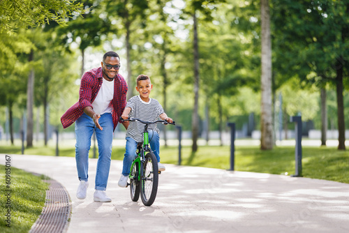 Happy ethnic family father teaches child son to ride bike in park .