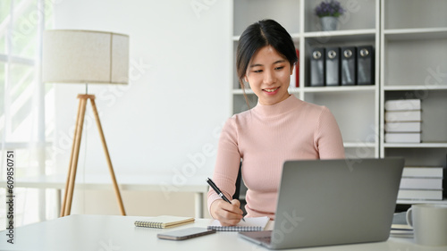 A beautiful Asian businesswoman focuses on her work, taking notes and using her laptop © bongkarn