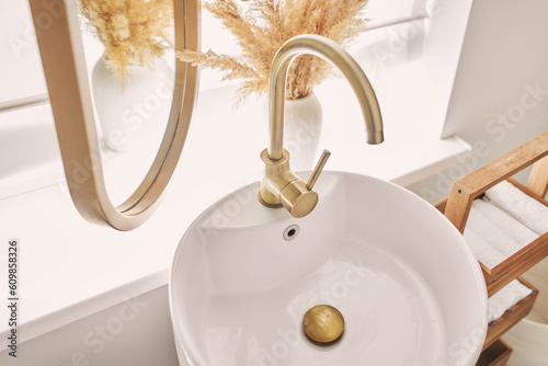 A beautiful sink with a golden faucet next to an oval mirror and a shelf with hand towels. Close-up of an elegant golden faucet in the bathroom sink next to stylish decorations.
