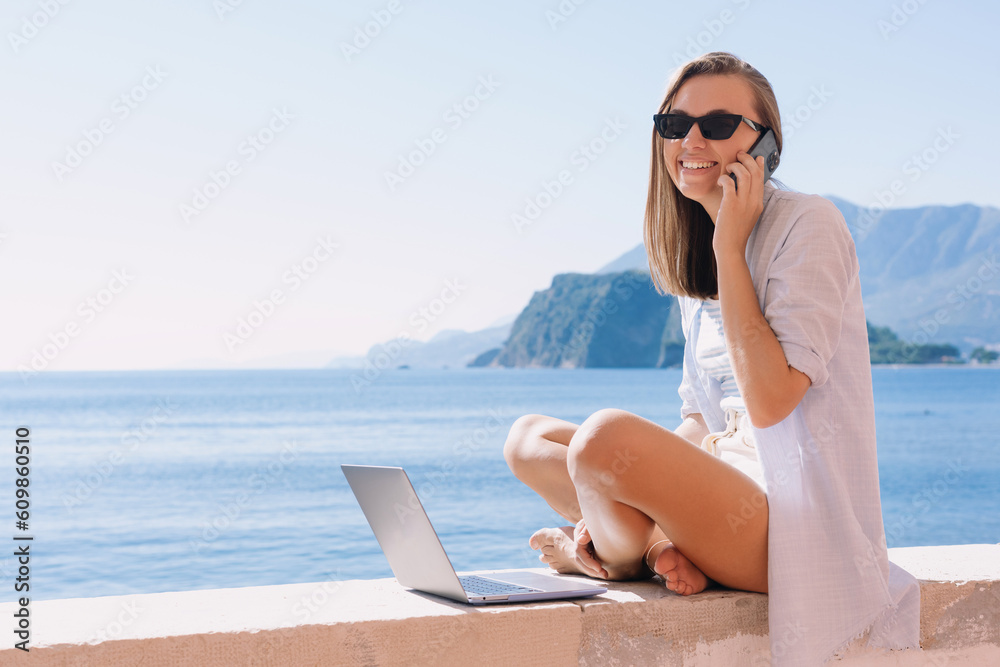 A smiling woman is sitting by the sea and talking on the phone. Freelancer in front of a laptop. Remote problem solving