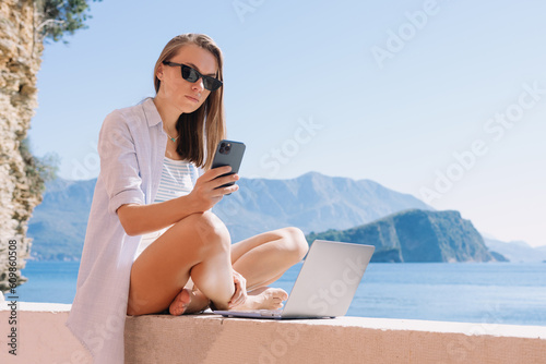 Work by the seashore. A young woman is typing on her phone, there is a laptop in front of her. Digital Nomad