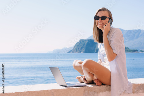 A smiling woman is sitting by the sea and talking on the phone. Freelancer in front of a laptop. Remote problem solving