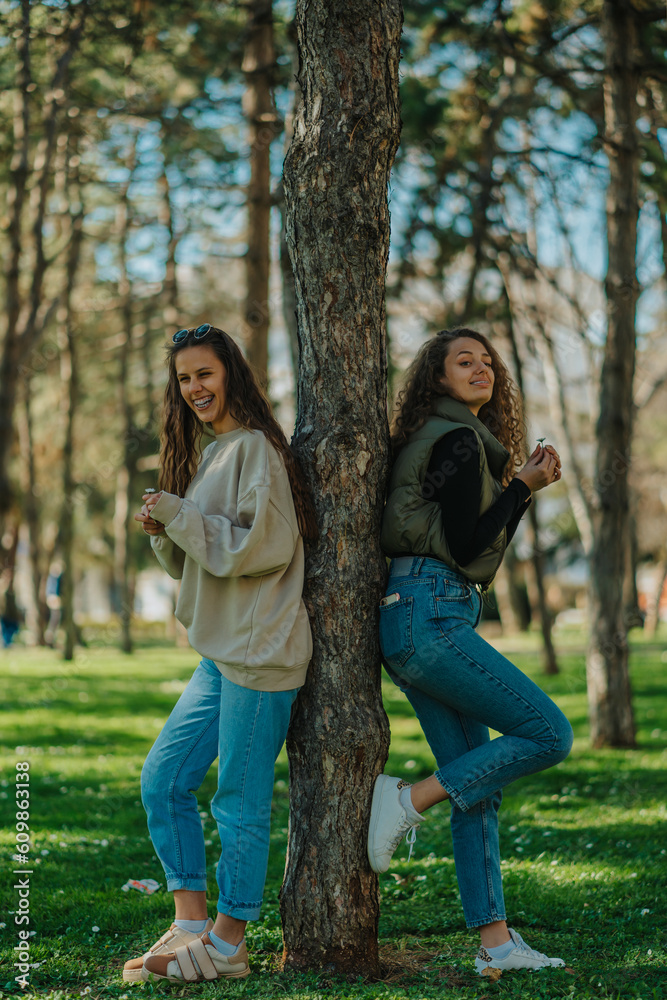 Long haired brunette girl burst out laughing while having a fun conversation with her lovely curly haired friend. They are both holding a flower in their hands and leaning against a tree in the park Long haired brunette girl burst out laughing while having a fun conversation with her lovely curly haired friend. They are both holding a flower in their hands and leaning against a tree in the park