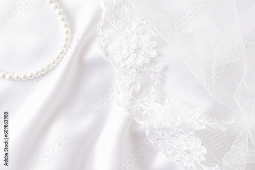 Chic wedding background for design. white satin fabric with soft waves, lace embroidered bridal veil and pearl necklace.