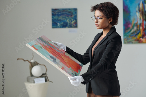 Young gallery owner in white gloves holding painting in her hands while working in gallery photo